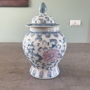 Może przedstawiać: Biały ceramiczny wazon z pokrywką, ozdobiony niebieskimi i różowymi wzorami kwiatowymi. Wazon ma pękaty korpus i zaokrągloną pokrywkę z małym, jasnoniebieskim guzikiem. Projekt obejmuje stylizowane liście i centralny kwiat.