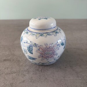 Puede incluir: Un pequeño tarro redondo de cerámica blanca con tapa. El tarro está decorado con un estampado floral en azul y rosa. La tapa tiene un diseño floral a juego. El tarro está sobre una superficie gris.