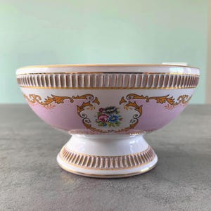 Könnte beinhalten: Eine rosa und weiße Keramikschale mit goldener Verzierung und Blumenmuster. Die Schale hat einen Sockel.
