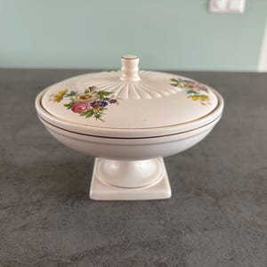 Op de afbeelding: Een witte keramische schaal met deksel op een voetstuk. Het deksel heeft een bloemenpatroon in roze, geel en paars. De schaal heeft een gouden rand en een kleine knop bovenop.