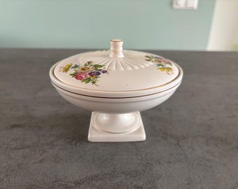 May include: A white ceramic lidded dish with a pedestal base. The lid is decorated with a floral pattern in pink, yellow, and purple. The dish has a gold trim and a small knob on top.