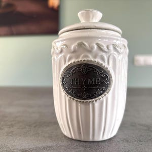 Może przedstawiać: Biały ceramiczny słoik z srebrną etykietą z napisem "THYME".