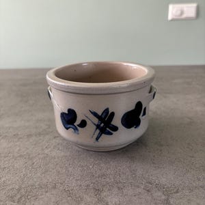 Op de afbeelding: Een kleine, witte keramische pot met blauwe beschilderde ontwerpen. De pot heeft twee kleine handvatten aan de zijkanten.