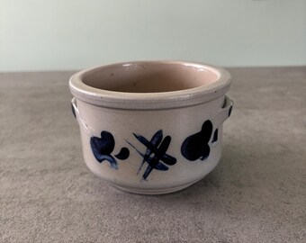 May include: A small, white ceramic pot with blue painted designs. The pot has two small handles on the sides.