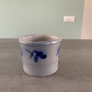 May include: A white ceramic jar with blue painted designs. The jar has a rounded bottom and a slightly flared top.