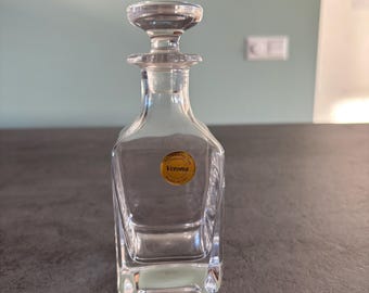 May include: A clear glass Verona decanter with a square base and a matching stopper. The decanter has a gold label. The stopper is round and fits snugly into the neck of the bottle. The decanter is empty and sits on a dark surface.