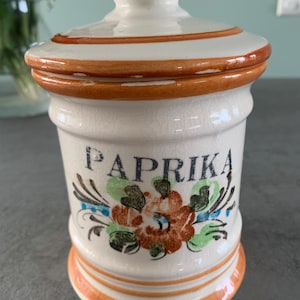 Op de afbeelding: Witte keramische paprikakruidenpot met deksel. De pot heeft bruine accenten en een bloemenontwerp met het woord "PAPRIKA" in zwarte letters. De pot is cilindrisch met een licht uitlopende basis.