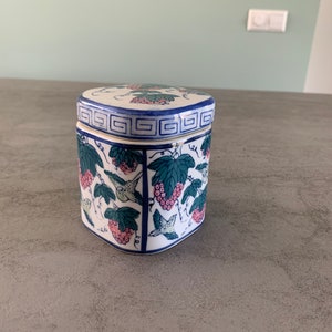 Pode incluir: Uma caixa de cerâmica branca com um design floral azul e verde. A caixa tem uma forma quadrada e uma tampa com um padrão geométrico.