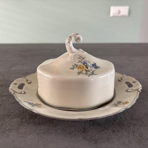 Peut inclure: Un beurrier en céramique blanche avec couvercle, orné de motifs floraux bleus et jaunes. Le plat repose sur une assiette assortie avec des poignées décoratives. Le couvercle a une poignée incurvée. Un article de cuisine classique.