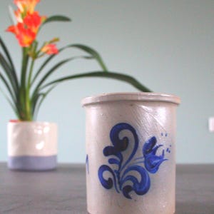 Op de afbeelding: Een witte keramische pot met een blauw bloemenmotief. De pot heeft een brede opening en een ronde bodem. Hij staat op een grijs oppervlak.