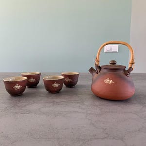 May include: A brown ceramic teapot with a bamboo handle and four matching teacups. The teapot and cups have a white design of a stylized bird.