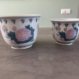 Op de afbeelding: Twee witte keramische bloempotten met een bloemenontwerp. De potten hebben roze en blauwe bloemen met groene bladeren. De randen hebben een blauw-wit geometrisch patroon. De potten zijn verschillend van grootte.