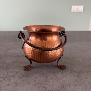 Op de afbeelding: Een kleine koperen pot met een gehamerde textuur en ijzeren handvatten. De pot heeft drie ijzeren poten en staat op een grijs oppervlak.