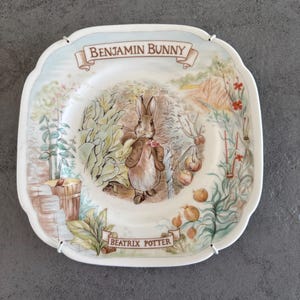 Op de afbeelding: Vierkante keramische plaat met een Benjamin Bunny-illustratie uit de verhalen van Beatrix Potter. De plaat heeft een geschulpte rand en een kleurrijke scène van een konijn in een tuin. De teksten "BENJAMIN BUNNY" en "BEATRIX POTTER" staan op de plaat.