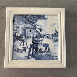 May include: A framed blue and white tile depicting a man crafting wooden shoes. The scene includes a rural setting with a house, windmill, and tools. The frame is a light, off-white color.