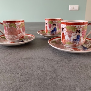 Op de afbeelding: Drie witte porseleinen theekopjes met rode en gouden bloemmotieven en afbeeldingen van vrouwen. De kopjes staan op bijpassende schoteltjes.