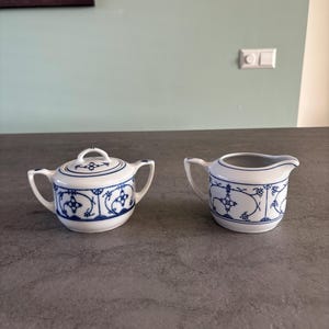 Puede incluir: Un azucarero de porcelana blanca con tapa y una jarrita para la crema, ambos adornados con un patrón floral azul. El azucarero tiene dos asas y la jarrita una sola asa y un pico. El conjunto está sobre una superficie gris.