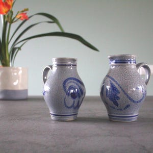 May include: Two grey ceramic jugs with blue floral designs. The jugs have a textured surface and a rounded shape.