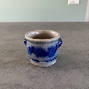 Pode incluir: Um pequeno pote de cerâmica cinza com esmalte azul e branco. O pote tem duas pequenas alças nas laterais e um design azul ondulado.