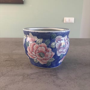 Peut inclure: Un petit pot en céramique de forme arrondie, présentant un fond bleu profond orné de grandes fleurs de pivoine peintes à la main en rose et blanc avec des feuilles vertes. Le bord est lisse et blanc, et l'intérieur a une finition blanche.
