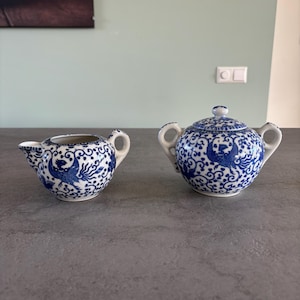 May include: A blue and white porcelain creamer and sugar bowl set. The set features a detailed blue phoenix and floral design on a white background. The sugar bowl has a lid and two handles. The creamer has a spout and a handle. The set is placed on a grey surface.