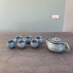 May include: A set of six light blue teacups and a matching teapot with a floral design. The teapot has a lid and a handle. The cups and teapot are on a gray surface.