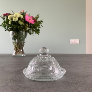 Könnte beinhalten: Eine klare Glasbutterdose mit einem gewölbten Deckel und einem strukturierten Muster. Die Dose steht auf einer grauen Arbeitsplatte.