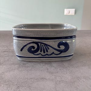 Puede incluir: Un cuenco rectangular de cerámica gris con una superficie texturizada y un diseño azul oscuro en forma de ola. El cuenco tiene una banda azul oscuro en la parte superior e inferior, y probablemente se utiliza para servir o exhibir.