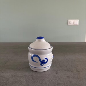 Op de afbeelding: Een kleine witte keramische pot met een blauw deksel en een blauw bloemenmotief. De pot staat op een grijs aanrecht.