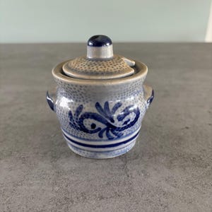 Op de afbeelding: Een kleine keramische suikerpot met een deksel. De kom is lichtblauw met een blauw bloemmotief en twee kleine handvatten. Het deksel heeft een kleine knop en een lepelhouder. De suikerpot staat op een grijze ondergrond.