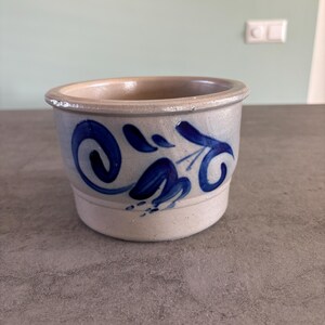 Op de afbeelding: Een kleine, ronde keramische pot met een lichtbeige interieur en een lichtblauwe buitenkant. De pot is versierd met een blauw bloemmotief. De pot staat op een grijze ondergrond.
