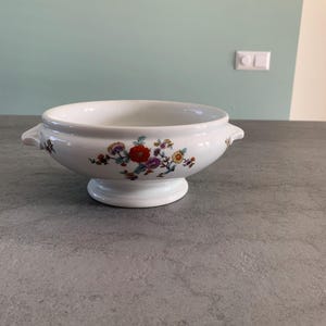 Op de afbeelding: Een witte keramische serveerschaal met twee handvatten en een bloemenmotief. De schaal heeft een voet en is versierd met een kleurrijk bloemenmotief met rode, gele en paarse bloemen.