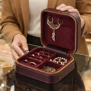 May include: A burgundy jewelry box with a gold zipper is open, revealing various pieces of jewelry. The interior is lined with a soft, dark red material. The jewelry includes rings, necklaces, and bracelets. The box is being held open by a person.