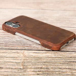 Puede incluir: Una funda de teléfono de cuero marrón con un acabado envejecido. La funda tiene un marco plateado y recortes para la cámara y el puerto de carga. La funda del teléfono está sobre una superficie de madera.