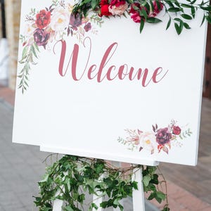 May include: A white wooden welcome sign with the word "Welcome" in a cursive font. The sign is decorated with floral arrangements in shades of red, pink, and burgundy. Green foliage adorns the top and base of the sign.