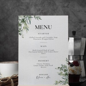 May include: A printed menu card with a wooden base, featuring a menu for a meal. The menu includes starters, main courses, and dessert options. The card is decorated with green botanical accents and the words "MENU" and "Olivia + Alex".