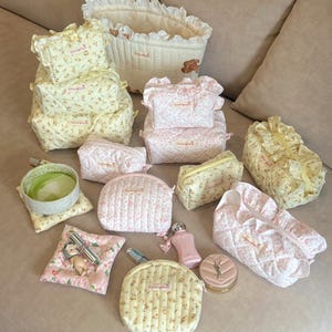 Puede incluir: Una colección de bolsas y estuches de tela acolchada con estampados florales en amarillo y rosa, con bordes de volantes y pequeñas etiquetas rectangulares. El conjunto incluye varias formas como estuches rectangulares, semicirculares y redondos.