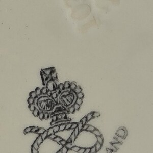 Puede incluir: Primer plano de una superficie color crema con un emblema en blanco y negro. El emblema presenta una corona sobre una letra "S" estilizada entrelazada con una cuerda, con las palabras "MADE IN ENGLAND" alrededor.