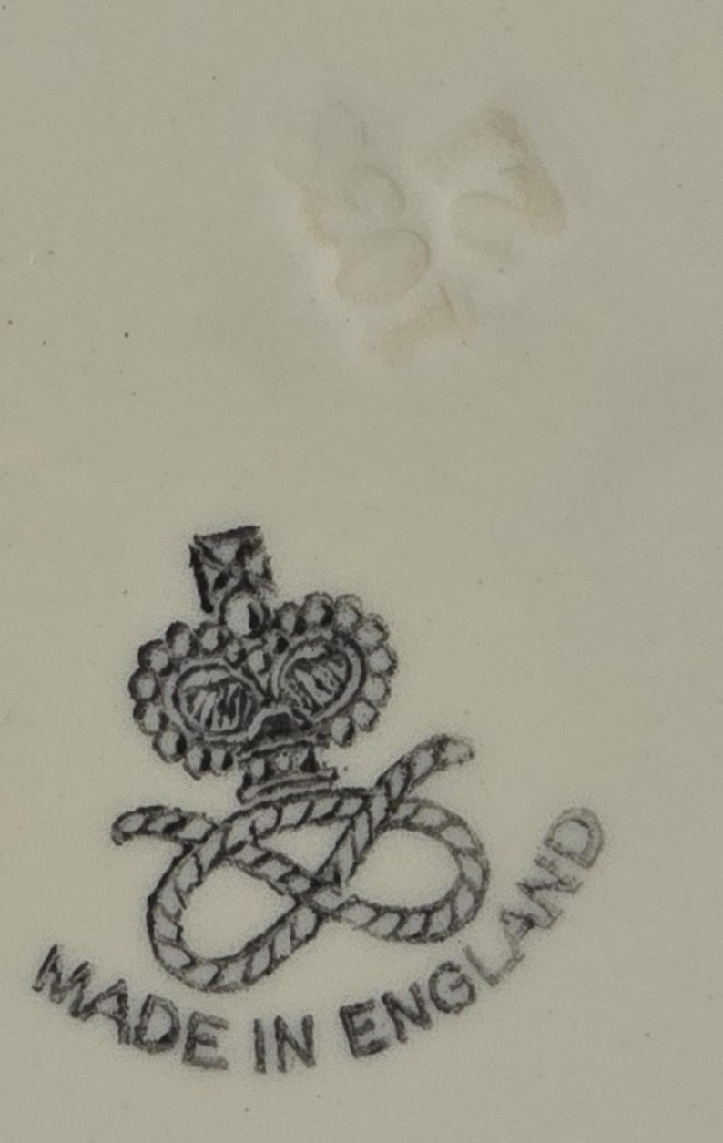 Puede incluir: Primer plano de una superficie color crema con un emblema en blanco y negro. El emblema presenta una corona sobre una letra "S" estilizada entrelazada con una cuerda, con las palabras "MADE IN ENGLAND" alrededor.