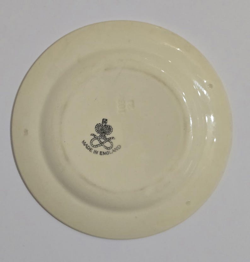 Puede incluir: Plato de cer&aacute;mica blanquecina con un borde ligeramente elevado. El centro presenta un emblema negro con una corona y la inscripci&oacute;n "MADE IN ENGLAND". El plato tiene un acabado brillante.