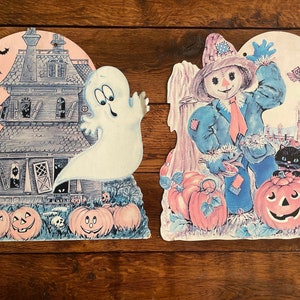 May include: Two vintage Halloween decorations. One features a haunted house with a ghost, bats, and pumpkins. The other depicts a scarecrow, pumpkins, a black cat, and an owl. Both have a pink moon background.
