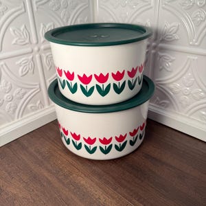 May include: Two white enamel bowls with green lids are stacked. Each bowl features a repeating pattern of red tulips with green stems and leaves. The bowls are on a dark wood surface, with a white tile background.
