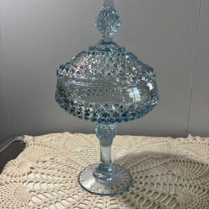 May include: A light blue, vintage-style glass candy dish with a lid. The dish has a pedestal base and a diamond-cut pattern. The lid is topped with a pointed finial. The dish is likely used for serving or displaying candies or small items.