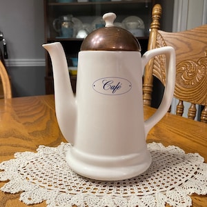 May include: White ceramic coffee pot with a copper lid and a blue oval label that says "Cafe". The pot is sitting on a white crocheted doily on a wooden table.
