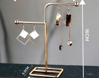 May include: Gold metal earring stand with three arms. The stand is 15 cm tall, 16 cm wide, and 7 cm in diameter. The stand has a square base.