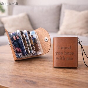 Peut inclure: Un petit album photo en cuir marron ouvert, affichant plusieurs photos. Un deuxième album en cuir marron fermé est à côté, avec les mots "I need you here with me" gravés sur le devant. L'album est sur une surface en bois.