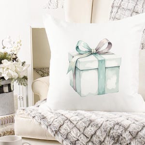 K&ouml;nnte beinhalten: Quadratisches wei&szlig;es Kissen mit einer Aquarellillustration einer Geschenkbox mit tealfarbenem Band und Schleife. Das Kissen liegt auf einem cremefarbenen Sofa mit einem Kunstfell&uuml;berwurf. Im Hintergrund ein Blumenarrangement.