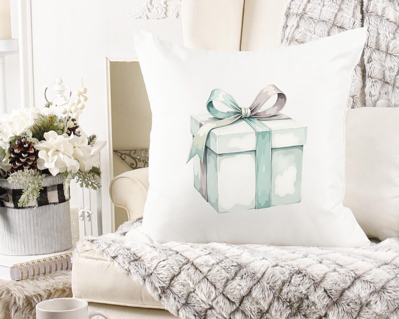 K&ouml;nnte beinhalten: Quadratisches wei&szlig;es Kissen mit einer Aquarellillustration einer Geschenkbox mit tealfarbenem Band und Schleife. Das Kissen liegt auf einem cremefarbenen Sofa mit einem Kunstfell&uuml;berwurf. Im Hintergrund ein Blumenarrangement.