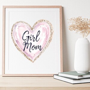 K&ouml;nnte beinhalten: Gerahmter Druck mit einem rosa Aquarellherz und goldfarbenem Glitzerbesatz. Die Worte "Girl Mom" sind in schwarzer Schrift geschrieben. Das Kunstwerk wird auf einer Holzoberfl&auml;che mit einer wei&szlig;en Vase und B&uuml;chern pr&auml;sentiert.