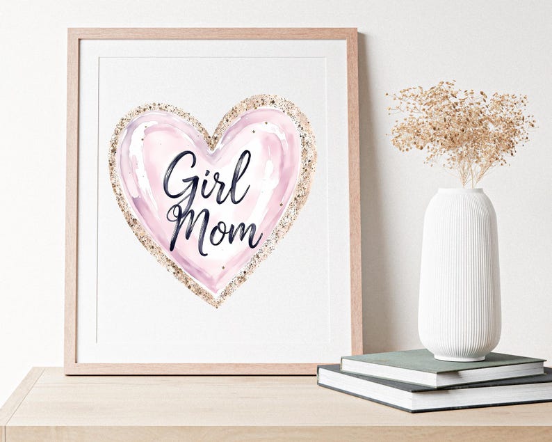 K&ouml;nnte beinhalten: Gerahmter Druck mit einem rosa Aquarellherz und goldfarbenem Glitzerbesatz. Die Worte "Girl Mom" sind in schwarzer Schrift geschrieben. Das Kunstwerk wird auf einer Holzoberfl&auml;che mit einer wei&szlig;en Vase und B&uuml;chern pr&auml;sentiert.